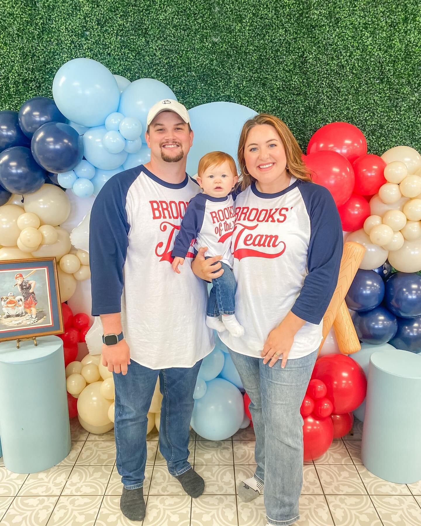 Kayla, her husband Brandon and their son Brooks in matching baseball shirts, in front of a blue and red balloon arch at Brooks' first birthday