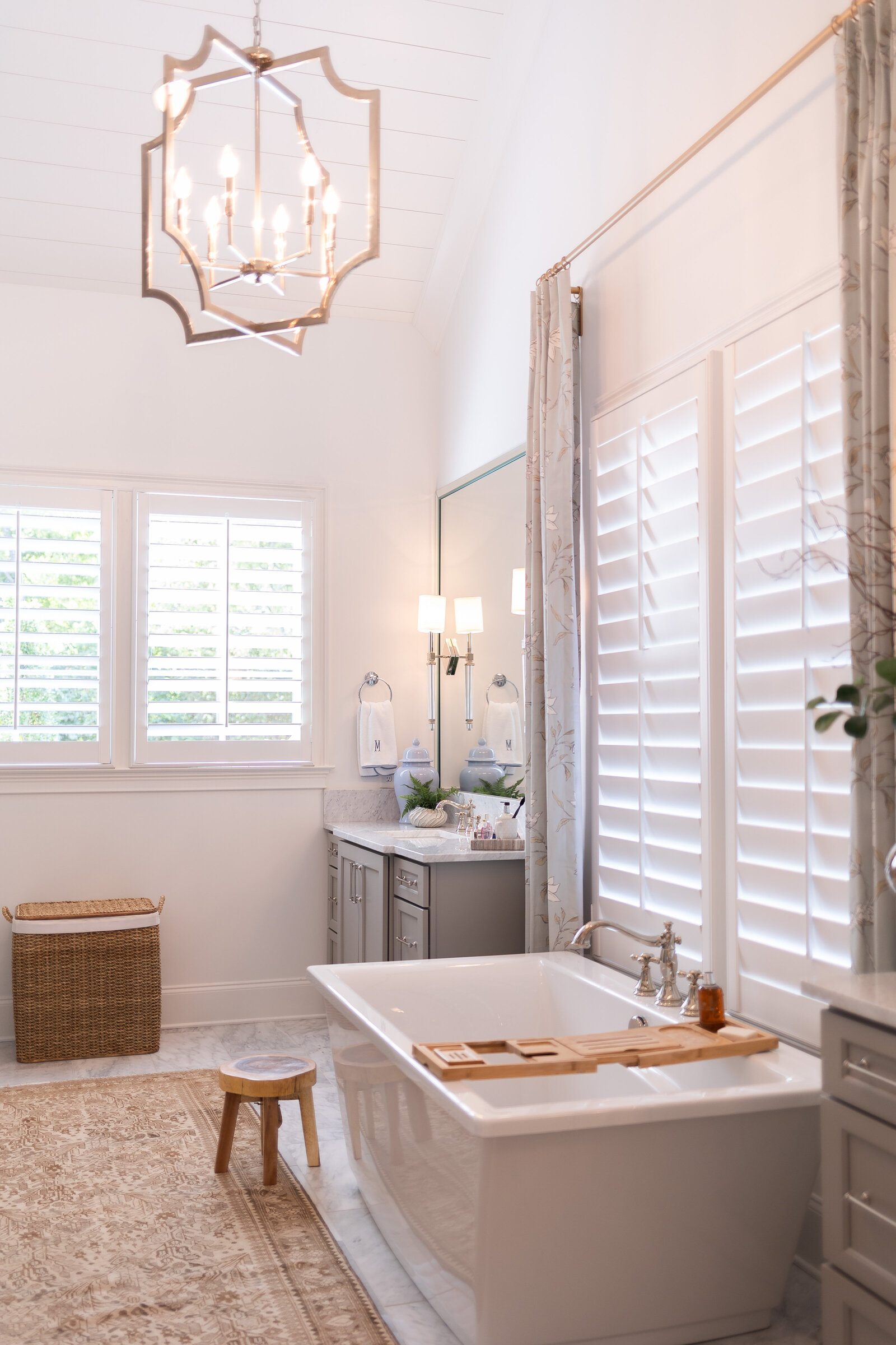 A primary bathroom with a lantern chandelier, a freestanding tub and wooden caddy, plantation shutters and a grey vanity