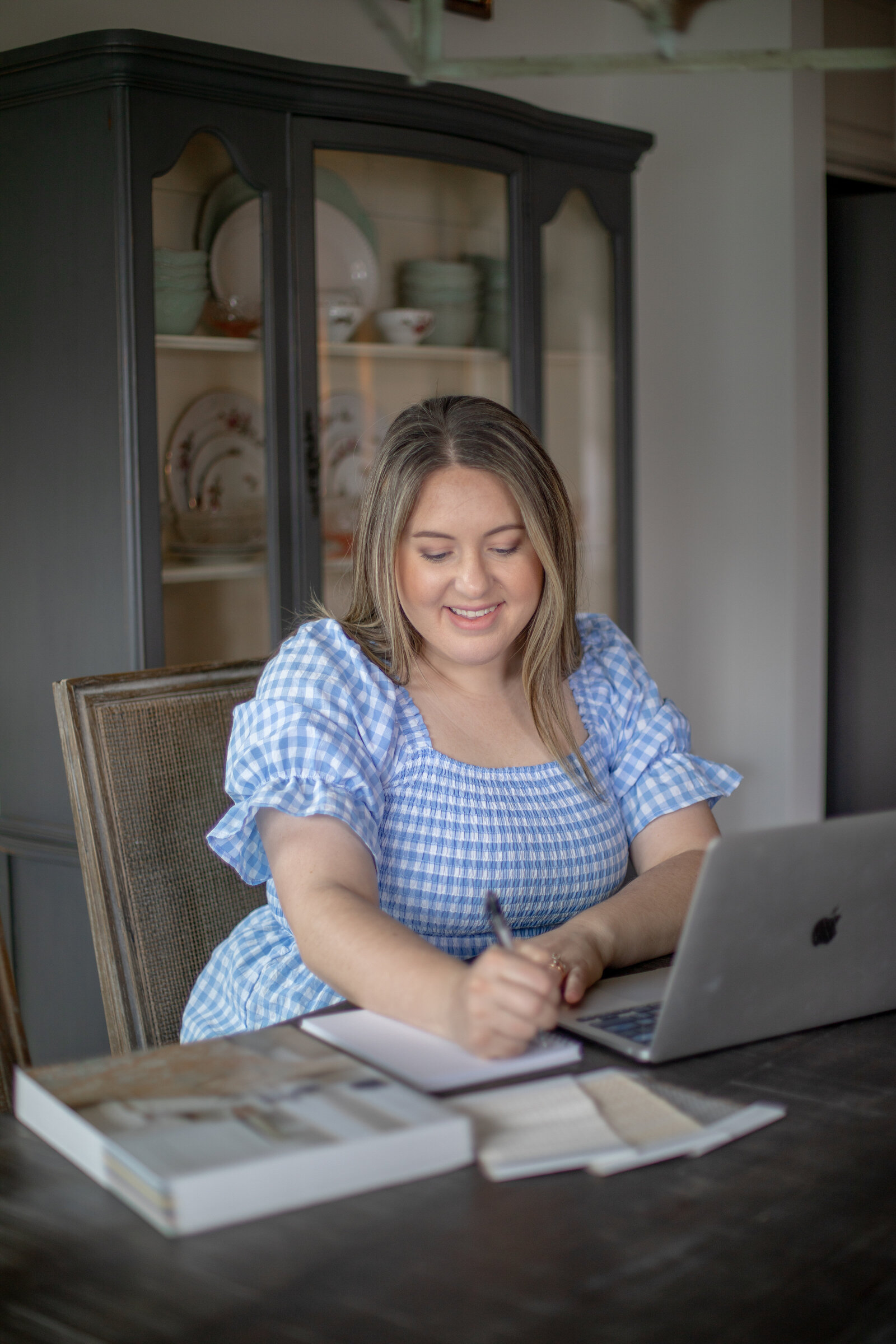 Kayla writing at a dark wood dining table beside an open laptop, fabric samples and a stack of design books