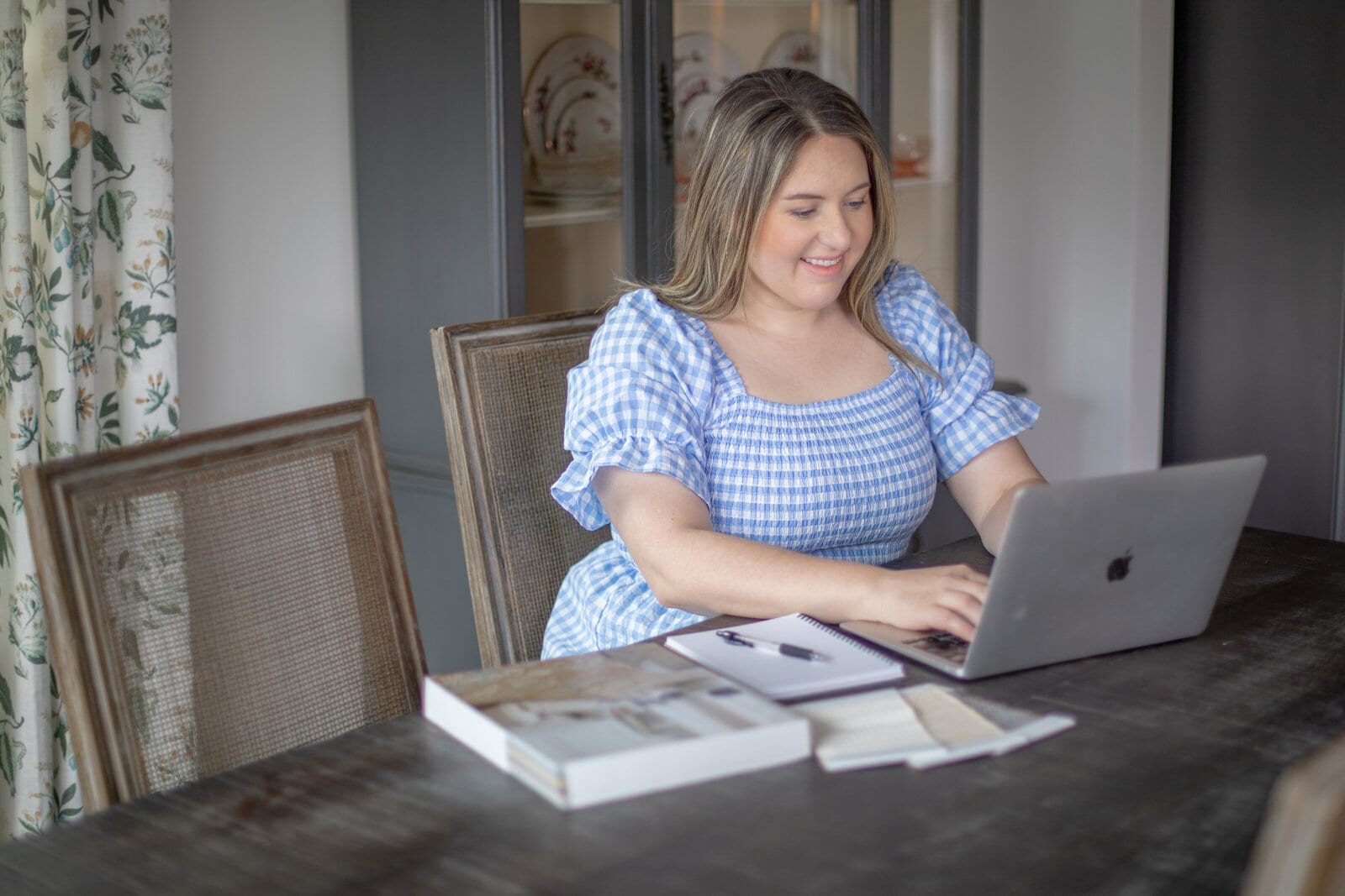Kayla in a blue gingham dress, typing on a laptop at a dark wood dining table, with a spiral notebook, design books and fabric samples beside her