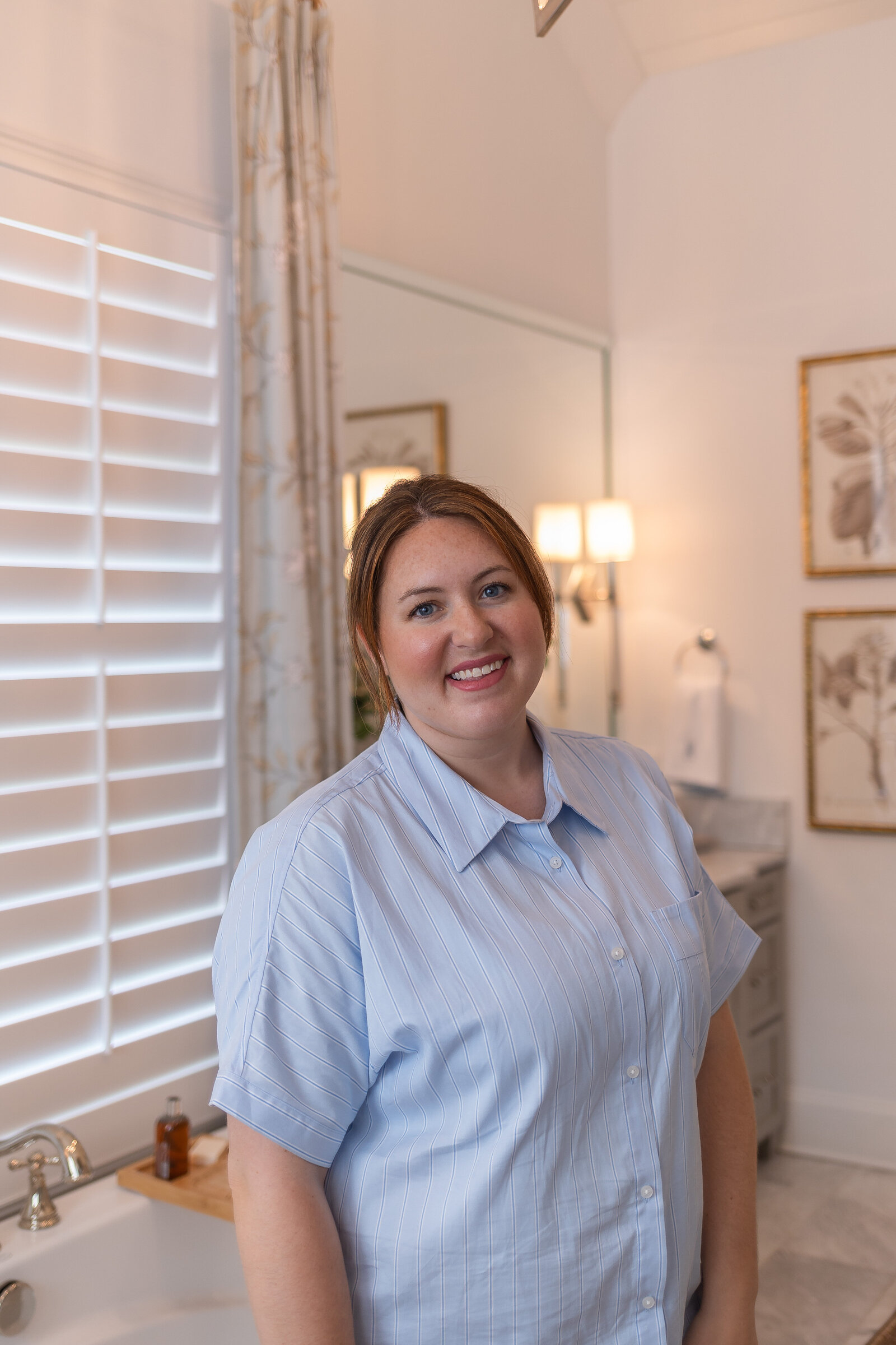 Kayla standing in a bathroom in a pale blue shirt, beside a freestanding tub, with plantation shutters and framed botanical prints