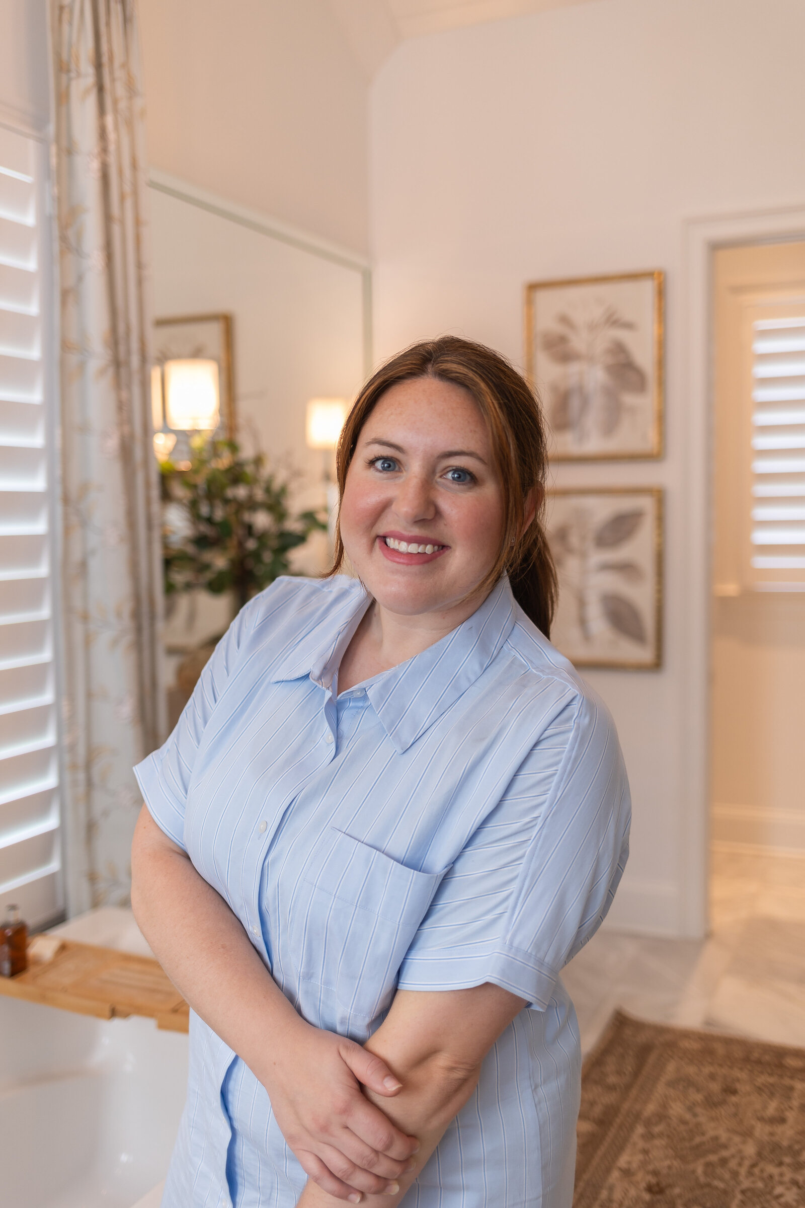 Kayla laughing in a bathroom in a pale blue shirt, with framed botanical prints on the wall behind her