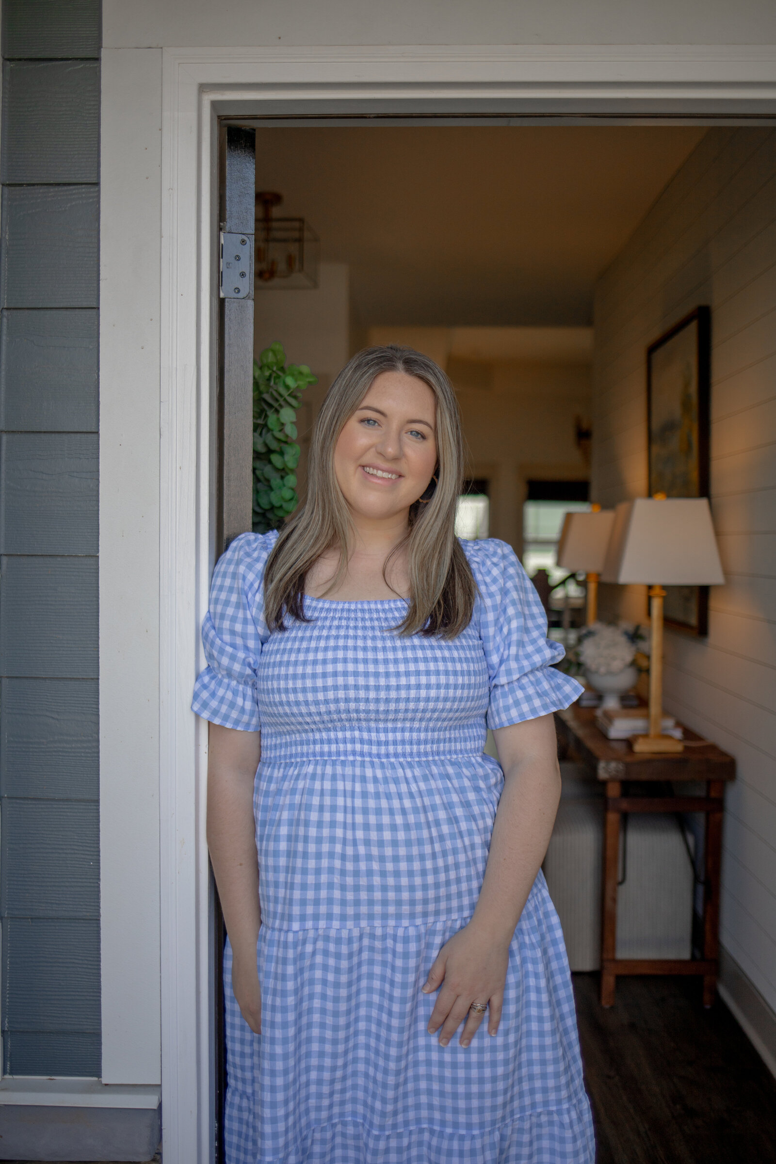 Kayla standing in the open doorway of a home, with a lamp-lit entry table behind her