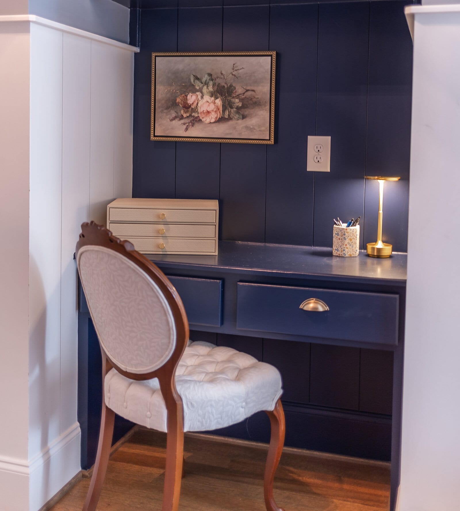 A navy built-in desk nook with a gold-framed floral still life, a cream jewellery chest, a brass lamp and an antique balloon-back chair