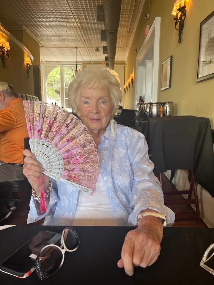 Gloria Dean seated at a restaurant table, holding up a pink lace hand fan