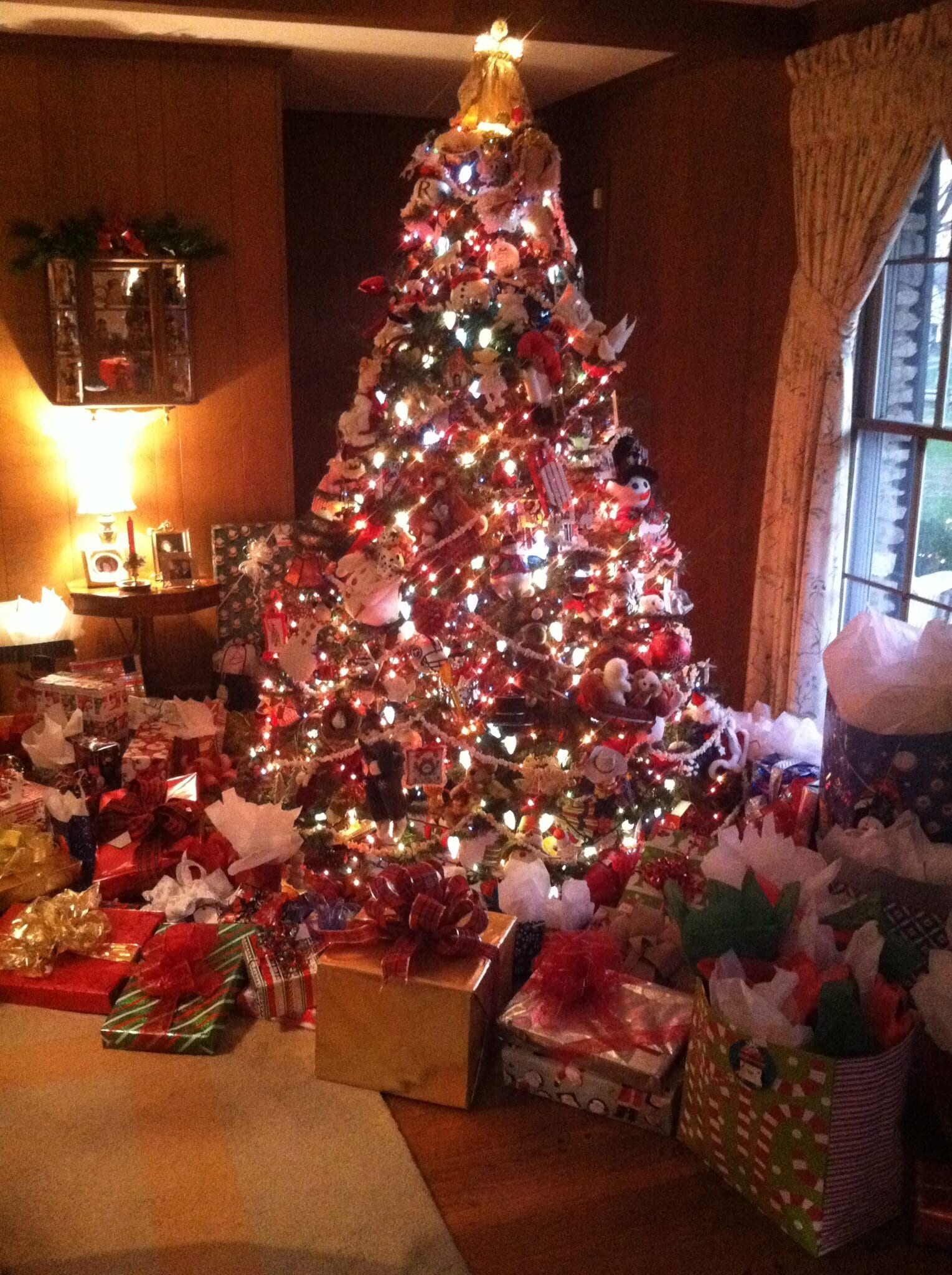 A Christmas tree crowded with red and white ornaments under a gold angel topper, with presents piled around it