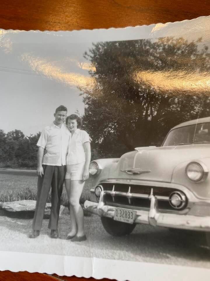A scallop-edged snapshot of Gloria Dean and her husband as a young couple, standing arm in arm beside a 1950s car