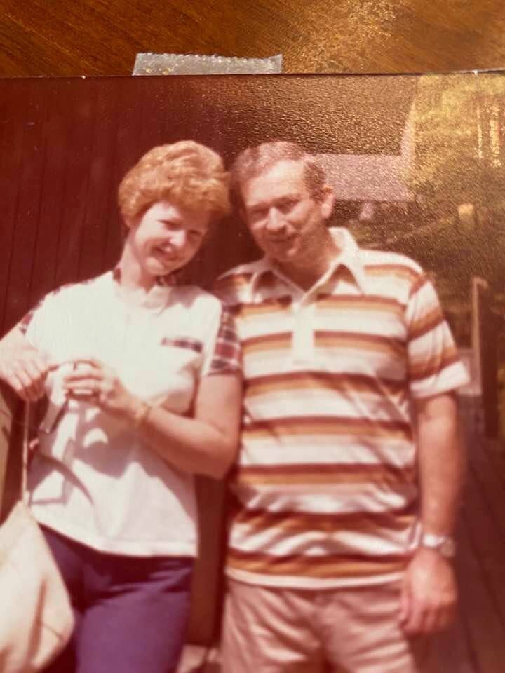 A warm-toned snapshot of Gloria Dean leaning against her husband in a striped polo shirt, outdoors in summer