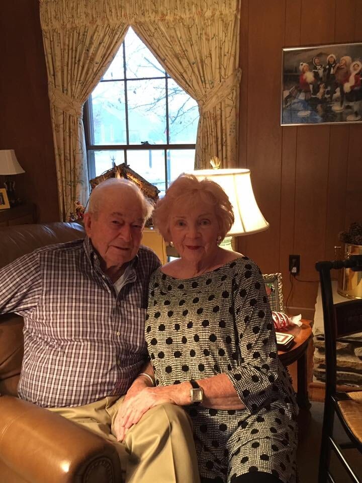 Gloria Dean and her husband in later life, sitting close together on a leather sofa, holding hands