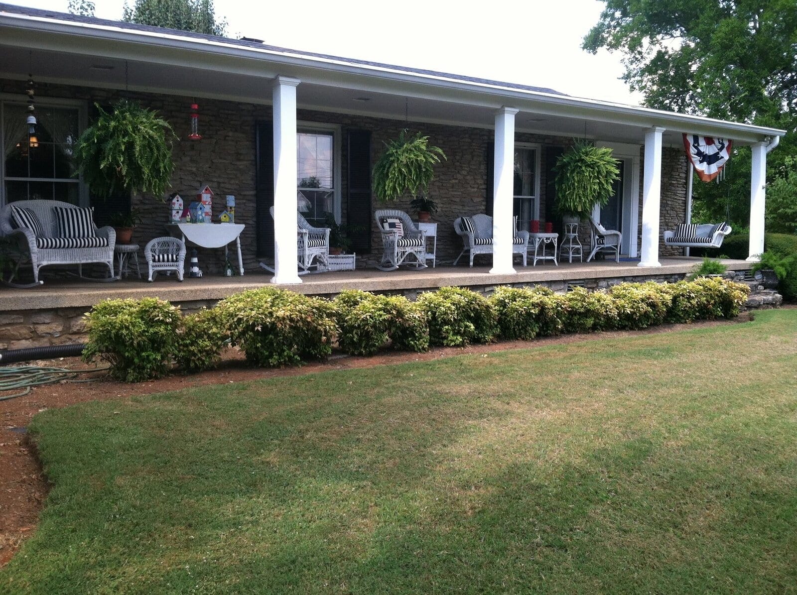 A long stone ranch house with a deep covered porch, white wicker furniture with striped cushions and hanging ferns